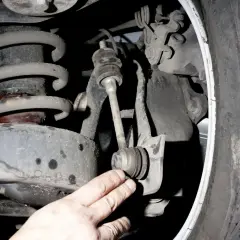 Dłoń wskazuje na zużyty stabilizator w zawieszeniu samochodu, obok sprężyny i opony.