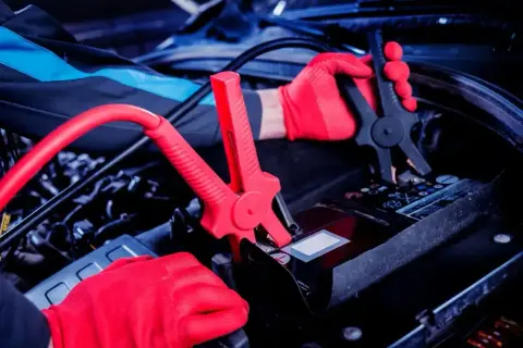 Ręce w czerwonych rękawiczkach podłączają kable rozruchowe do akumulatora. Czy nowy akumulator trzeba ładować? To pytanie często zadawane przez kierowców.