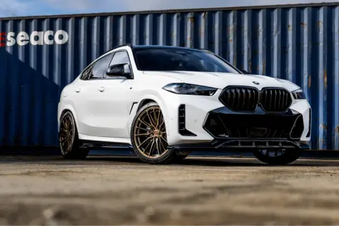 Białe BMW X6 z brązowymi felgami, na których zamontowano opony Imperial, prezentuje się imponująco na tle niebieskiego kontenera.