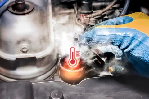 Rękawica mechanika sprawdza zbiorniczek wyrównawczy, z którego wydobywa się para. Widoczny symbol termometru wskazuje na **wysoką temperaturę płynu chłodniczego**.