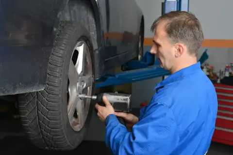 Mechanik w niebieskim kombinezonie dokręca śruby kół aluminiowych przy podniesionym samochodzie.