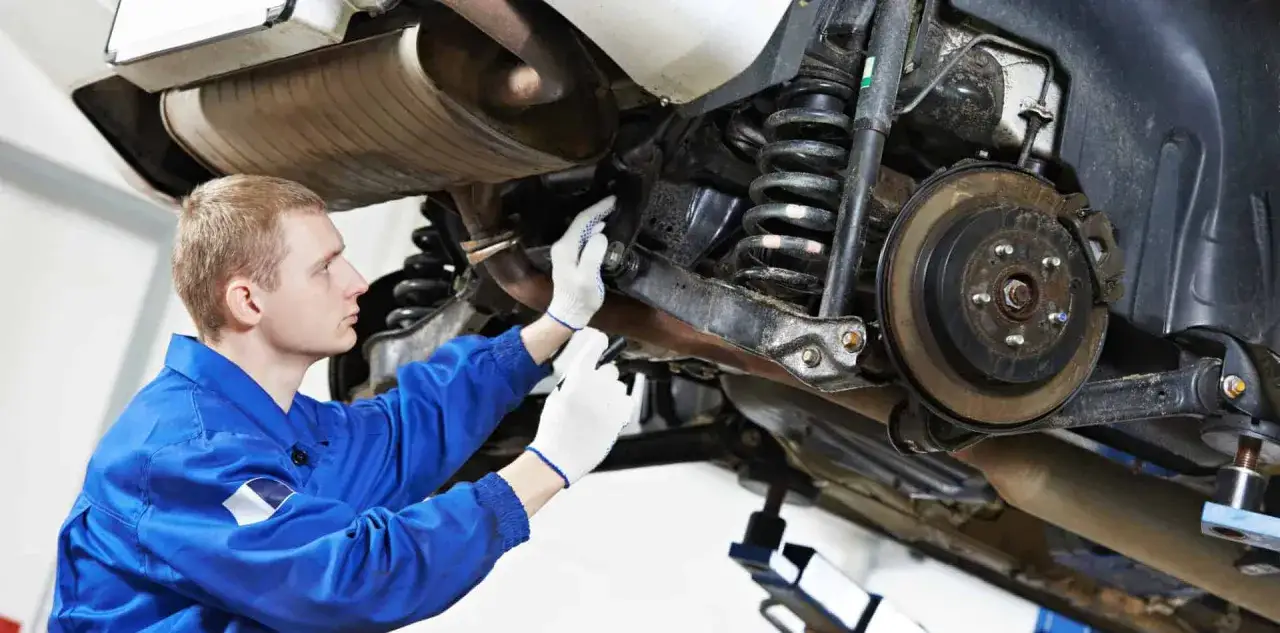 Mechanik w niebieskim kombinezonie sprawdza zawieszenie samochodu, szukając przyczyny głuchego stukania podczas jazdy.
